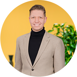 Een man met kort haar in een beige blazer en zwarte coltrui, die glimlachend voor een gele achtergrond met groene plant staat.