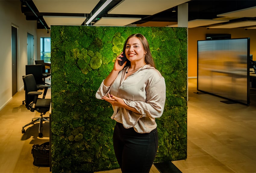 Vrouw met lang bruin haar voert een telefoongesprek in een modern kantoor, staand voor een groene moswand en glimlachend naar de camera.