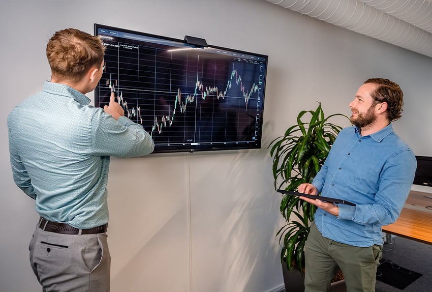 Twee mannen bespreken een grafiek op een groot scherm in een kantoorruimte.