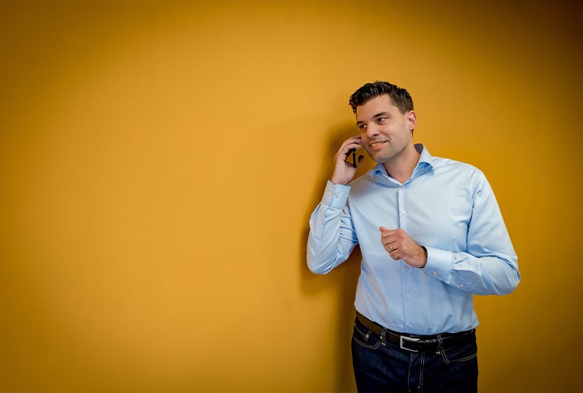 Man in een lichtblauw overhemd belt glimlachend met zijn mobiele telefoon tegen een gele achtergrond.