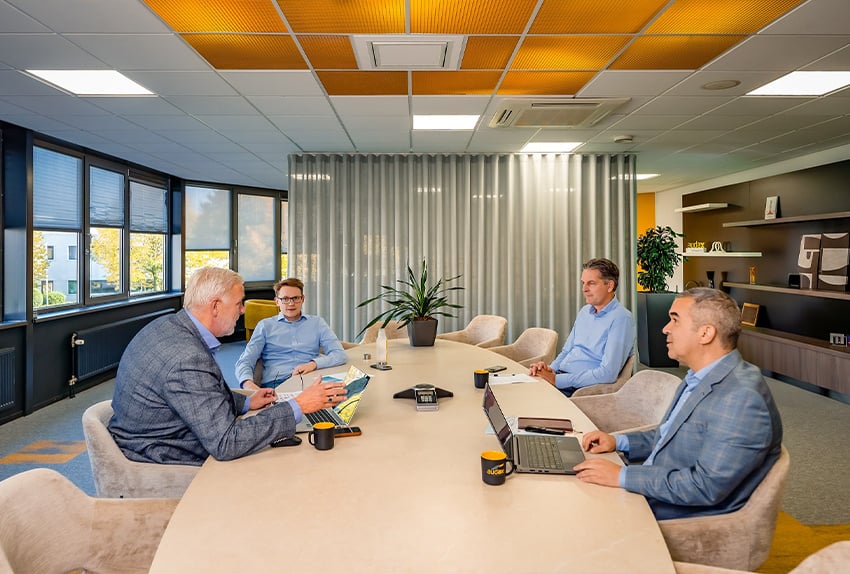 Vier mannen in overleg tijdens een zakelijke vergadering in een modern kantoor met grote ramen, laptops op tafel en een plant in het midden van de vergadertafel.