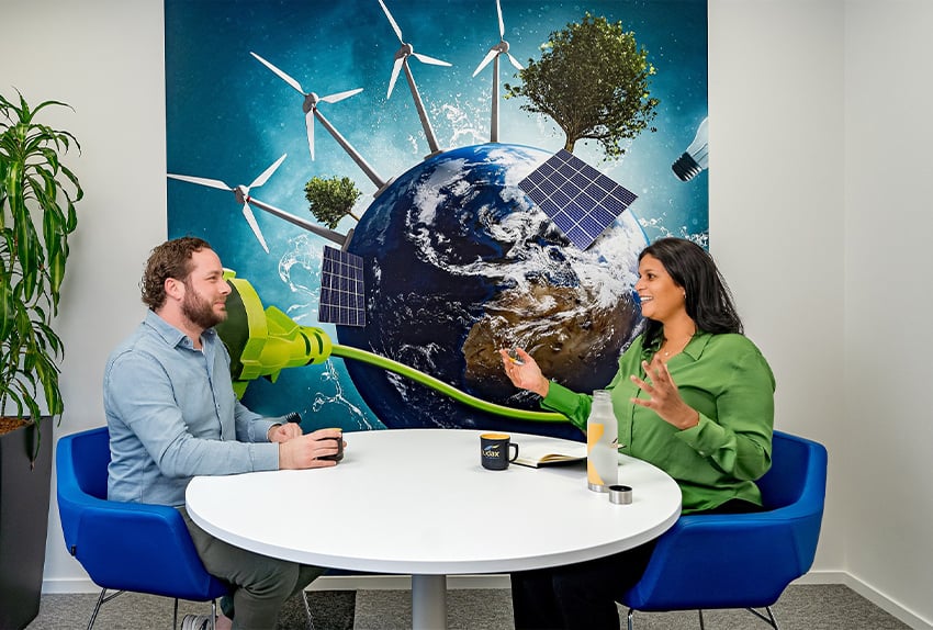 Twee collega’s in gesprek aan een ronde tafel in een kantoor, met op de achtergrond een kleurrijke muurschildering van de aarde omringd door windmolens, zonnepanelen, bomen en een groene stekker als symbool voor duurzaamheid en groene energie.