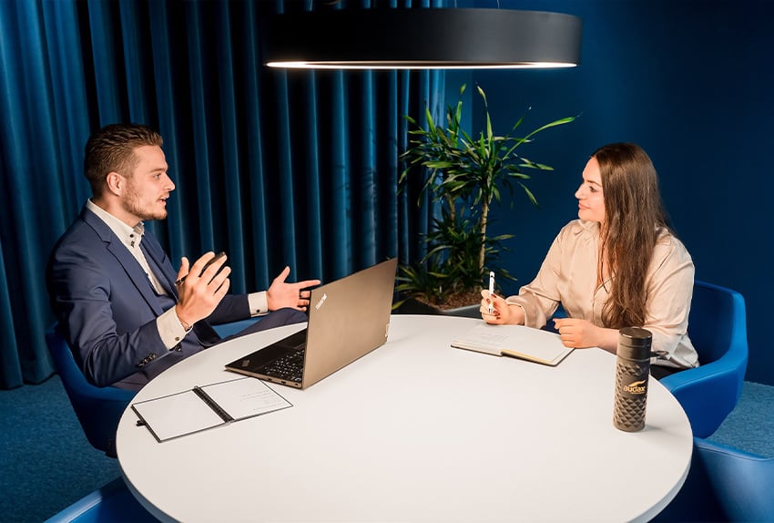 Twee collega’s in gesprek aan een ronde tafel in een modern, blauwgestyled kantoor, met een open laptop en notities voor zich.