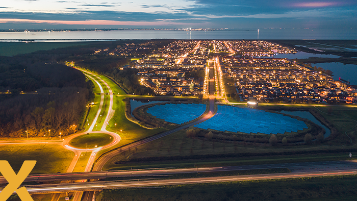 Luchtfoto van een verlichte stad bij schemering met zonnepark en snelweg op de voorgrond.