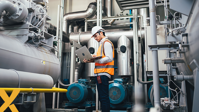 Energiespecialist met veiligheidshesje en helm inspecteert industriële installaties terwijl hij werkt op een laptop.