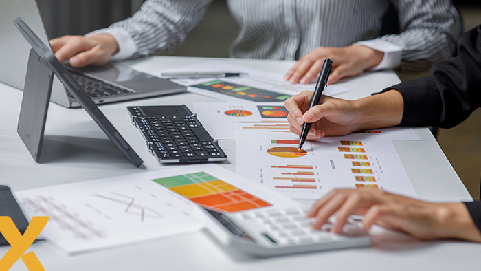 Twee personen zitten aan tafel en analyseren grafieken en cijfers met laptop en rekenmachine.