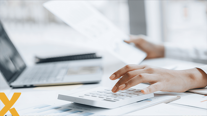 Handen van een persoon die met een rekenmachine en documenten werkt, met een laptop op de achtergrond — zakelijke administratie of energiekostenberekening.
