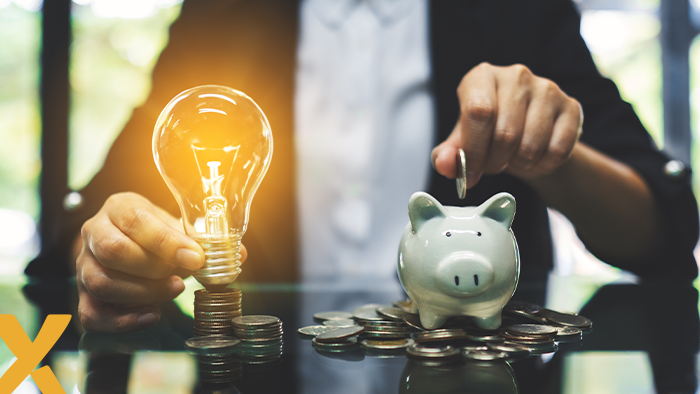 Een hand plaatsteen munt in spaarvarken, welke naast een brandende gloeilamp en stapels munten staat.