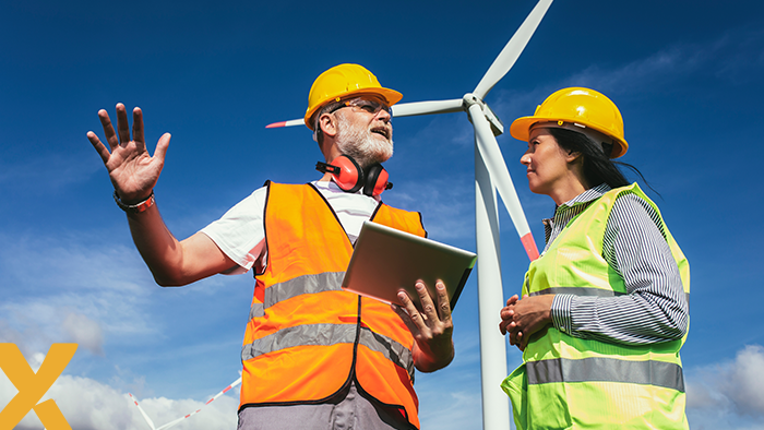 Technici met veiligheidshelmen en tablets bespreken werkzaamheden bij een windturbine op een zonnige dag, symbool voor duurzame energieproductie.