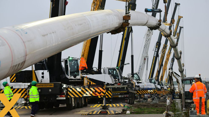 Bouwplaats waar meerdere kranen een grote pijpleiding optillen, met arbeiders in veiligheidskleding op de voorgrond.