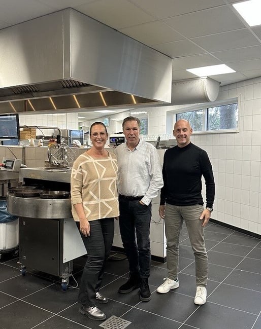 Drie personen poseren samen in een professionele keuken met witte betegelde muren en een grote roestvrijstalen afzuigkap.