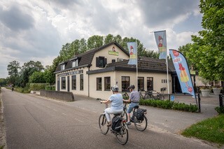 Twee fietsers die langs het licht ecru gekleurd pand van Koeckers met kleurrijke vlaggen fietsen op een rustige weg, omringd door groene bomen onder een bewolkte lucht.