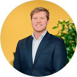 Portret van een man met blond haar, in donkerblauw colbert tegen een gele achtergrond met een groene plant op de achtergrond.